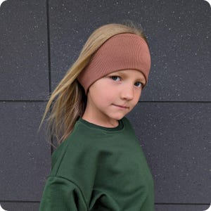 Op de afbeelding: Een jong persoon draagt een geribbelde, stoffig roze hoofdband en een donkergroene trui. De hoofdband bedekt het voorhoofd en de oren. De persoon heeft lang, blond haar en kijkt in de camera.