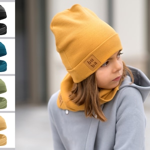 May include: A yellow knit beanie with a brown leather patch that says "bu ba" and a matching yellow knit scarf. The beanie and scarf are worn by a child with long brown hair.
