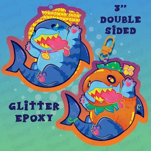May include: Double-sided, 3-inch glitter epoxy shark keychain. One shark is blue with a pink tongue and the name "Jeff" on a tag. The other shark is orange with a green and yellow floral design. The keychains have a gold-colored clasp.