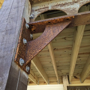 May include: Close-up of a weathered wooden post with a rusty, textured metal bracket bolted to it. The bracket supports wooden beams, part of a deck or porch structure. The wood has a natural grain, and the metal shows signs of age.