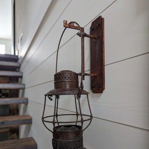 May include: A rusty metal lantern with a wire cage and a decorative top hangs from a rusty metal bracket on a white wall.