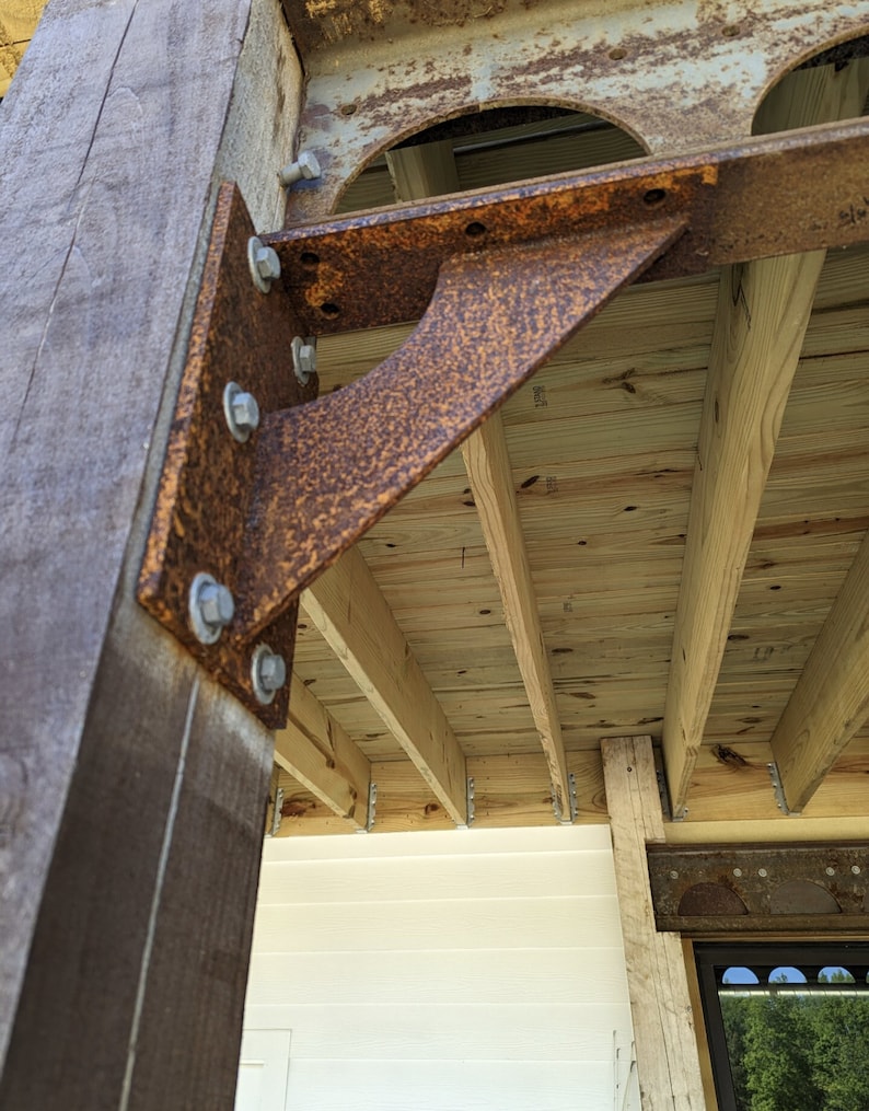 Pu&ograve; includere: Primo piano di un palo di legno usurato con una staffa metallica arrugginita e strutturata imbullonata. La staffa supporta travi di legno, parte di una struttura di terrazza o portico. Il legno ha una venatura naturale e il metallo mostra segni di et&agrave;.
