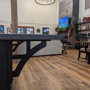 May include: A close-up of a black metal bracket supporting a countertop. The bracket has a decorative design and is attached to a wooden beam. The countertop is made of concrete and has a smooth, gray surface.