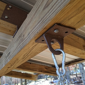 May include: A rusty metal swing set bracket attached to a wooden beam with a silver carabiner and chain hanging from it.