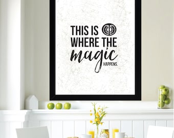 May include: A black framed print with white background and black text that reads "This is where the magic happens."  The print is hanging on a white wall above a white table with two white chairs. There are green apples on the table and a vase with yellow flowers.
