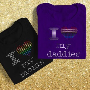 Pode incluir: Duas camisetas pretas e roxas com desenhos de corações arco-íris e o texto "Amo minhas mães" e "Amo meus pais".