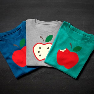 May include: Three t-shirts with different apple designs. The first t-shirt is blue with a red apple. The second t-shirt is gray with a green apple that is cut in half. The third t-shirt is teal with a red apple that has a bite taken out of it.