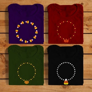May include: Four folded t-shirts with different autumn-themed designs. The shirts are purple, red, green, and black. Each shirt features a circle of small images, such as candy corn, leaves, and pumpkins, with a larger image in the centre.