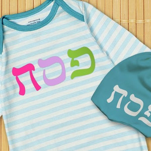 May include: A blue and white striped baby onesie with the Hebrew word "Sada" in pink, purple, and green letters. A matching blue baby hat with the same Hebrew word in white letters.