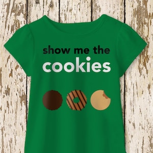 Op de afbeelding: Groen T-shirt met de tekst "show me the cookies" in zwart en wit. Onder de tekst staan drie koekjes.