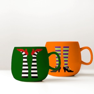May include: Two ceramic mugs with Halloween-themed designs. One mug is green with a black and white striped witch's leg design and red shoes. The other mug is orange with a purple and green striped witch's leg design and black shoes.