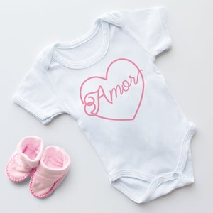 Peut inclure: Une grenouillère blanche pour bébé avec un motif de cœur rose qui dit "Amor" à l'intérieur. La grenouillère est accompagnée de chaussons roses pour bébé.