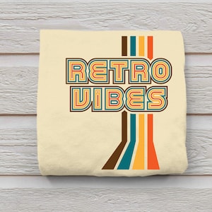 May include: A beige t-shirt with the text "Retro Vibes" in a retro font with colorful stripes. The text is in a multi-colored font with brown, teal, yellow, and orange stripes.