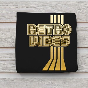 May include: Black t-shirt with gold foil lettering that reads "Retro Vibes" in a retro font style. The lettering is outlined with a thin black line. There are five vertical gold lines to the right of the text.