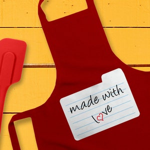 May include: Red kitchen apron with a white index card attached that says "made with love" with a red heart drawn next to the word "love". A red silicone spatula is on the left side of the image.