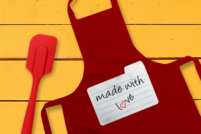 May include: Red kitchen apron with a white index card attached that says "made with love" with a red heart drawn next to the word "love". A red silicone spatula is on the left side of the image.