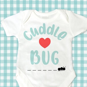 Puede incluir: Un body blanco para bebé con el texto "cuddle bug" en letras azul turquesa con un corazón rojo. Hay un gráfico de hormiga negra debajo del texto.