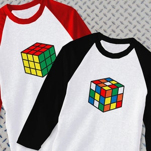 May include: Two baseball style t-shirts with a colorful Rubik's Cube graphic on the front. One shirt has red and black sleeves, the other has black and white sleeves.