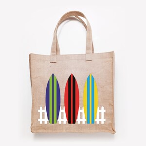 May include: A brown burlap tote bag with three colorful surfboards and a white picket fence design. The surfboards are blue, red, and yellow with green, black, and blue stripes.