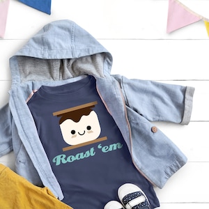 Könnte beinhalten: Ein marineblaues T-Shirt mit einem Motiv eines lächelnden Marshmallows mit Schokolade und Graham-Cracker obenauf. Der Text "Roast 'em" ist unter dem Motiv aufgedruckt.