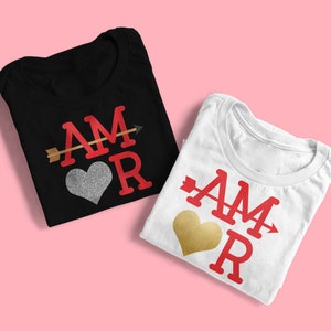 May include: Two folded t-shirts with the word "AMOR" printed on them in red letters. One shirt is black with a silver glitter heart and the other is white with a gold glitter heart. Both shirts have a gold arrow through the word.