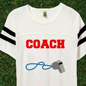 May include: White t-shirt with black and white striped sleeves. The shirt has the word "COACH" printed in red letters and a whistle with a blue string.