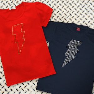 May include: Two t-shirts with a lightning bolt design made of rhinestones. One shirt is red with gold rhinestones and the other is navy blue with silver rhinestones.