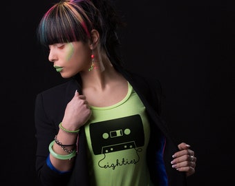 May include: A woman with colourful hair is wearing a green t-shirt with a black cassette tape graphic and the text "eighties". She is wearing a black blazer and has green makeup on her face.