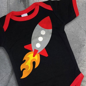 May include: A black baby onesie with a red trim around the neckline and sleeves. The onesie has a gray rocket ship with red and orange flames printed on the front.