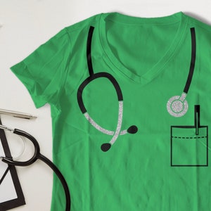May include: A green t-shirt with a black and silver glitter stethoscope design. The shirt has a pocket with a pen in it.
