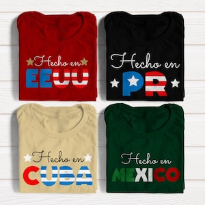 May include: Four folded t-shirts with different colors and designs. The shirts feature the text "Hecho en" followed by the name of a country in Spanish, with the letters in the country name styled in the colors of the country's flag. The shirts are red, black, beige, and green.