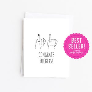 May include: A white greeting card featuring a black line drawing of two hands with middle fingers raised, each wearing a ring. The text below the hands reads CONGRATS FUCKERS! in a simple black font. The card is presented on a plain white surface.