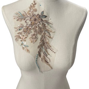 May include: A delicate floral applique with sequins and beads in shades of beige, brown, and teal. The applique is on a sheer mesh fabric and is perfect for adding a touch of elegance to clothing or crafts.