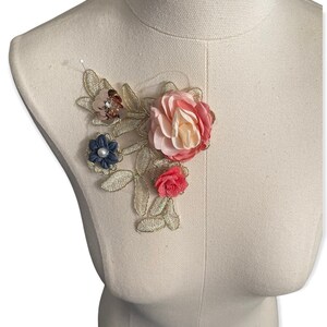 May include: A floral applique featuring fabric flowers in shades of pink, red, and blue, accented with gold-colored embroidered leaves. The applique is attached to a mannequin, showcasing its design and potential use for embellishment.