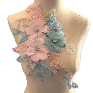 May include: A floral applique featuring three flowers in shades of pink and blue, with delicate leaves and accents. The applique is displayed on a mannequin, showcasing its design and potential use in fashion or craft projects.
