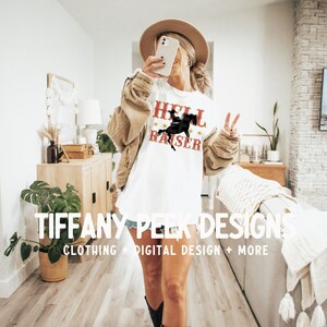 May include: A white t-shirt featuring the red text HELL RAISER and a black silhouette of a person riding a horse. The shirt is worn with a tan corduroy jacket, denim shorts, a tan hat, and black cowboy boots. The text TIFFANY PEEK DESIGNS is visible.