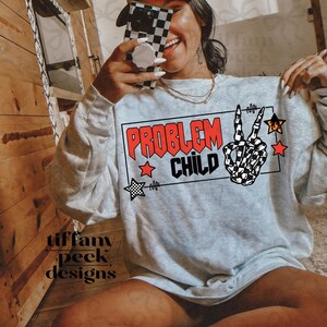 Könnte beinhalten: Hellgraues Sweatshirt mit dem Schriftzug "PROBLEM CHILD" in Rot und Schwarz, mit einer Skeletthand, die ein Friedenszeichen zeigt. Das Design enthält rote und schwarze Sterne. Der Markenname "Tiffany Peek Designs" ist sichtbar.