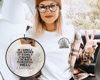 Può includere: Una t-shirt bianca con una piccola grafica circolare sul petto raffigurante una mano di scheletro che fa il segno di pace e il testo 'YOU CAN BET YOUR ASS I'M GONNA BE JUST FINE'. Un inserto circolare più grande mostra il testo sul retro 'IF I SMELL LIKE SMOKE IT'S ONLY CAUSE I'VE BEEN THROUGH HELL & BACK AGAIN'.