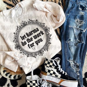 May include: A cream-colored crewneck sweatshirt featuring a black circular ornate frame graphic with the text let karma do the work for you, paired with distressed blue denim jeans and black and white checkered high-top sneakers.