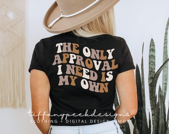 Può includere: Una t-shirt nera a maniche corte con il testo "THE ONLY APPROVAL I NEED IS MY OWN" in un carattere ondulato multicolore con tonalità marrone, beige e crema. La maglietta è indossata con un cappello a tesa larga beige e jeans azzurri.