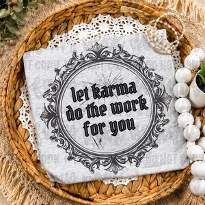 May include: A light gray t-shirt featuring a black circular frame with ornate scrollwork details. Inside the frame, the text reads, let karma do the work for you, with a cracked glass effect overlaying the lettering and the frame design.