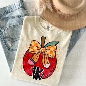 May include: A cream-colored t-shirt featuring a graphic of a red apple with a yellow and orange checkered bow and a pencil tucked behind it. The text 'tk' is printed in black below the apple. The shirt has a Comfort Colors brand neck label.