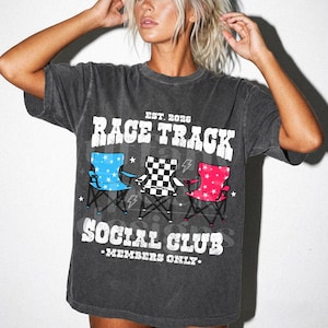 以下が含まれることがあります： 「RACE TRACK SOCIAL CLUB - MEMBERS ONLY」と書かれたダークグレーのTシャツ。青、白黒チェック、ピンクの折りたたみ椅子が3つ、星と稲妻のグラフィックが描かれています。
