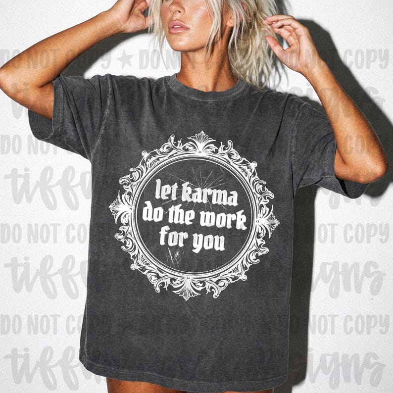 May include: A charcoal grey short-sleeve t-shirt featuring a white graphic print of an ornate, circular filigree frame. Inside the frame, the text reads, "let karma do the work for you" in a bold, stylized font. The shirt has a washed-out finish.
