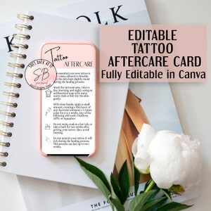 Op de afbeelding: Een roze smartphone met tattoo nazorginstructies, naast een kaart met de tekst "Editable Tattoo Aftercare Card Fully Editable in Canva". De afbeelding bevat een witte bloem, groene bladeren en een spiraalgebonden notitieboek.