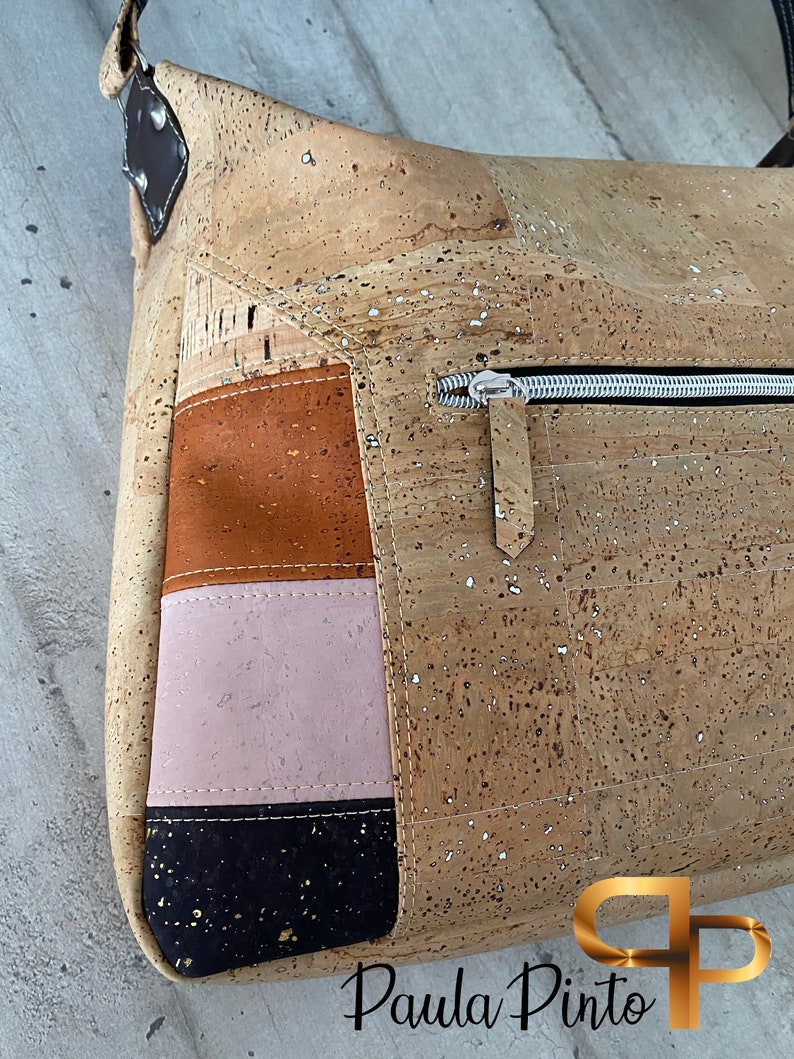 May include: A cork handbag with a zipped pocket and a patchwork design featuring brown, pink, and black fabric. The bag has a gold logo that says "Paula Pinto".