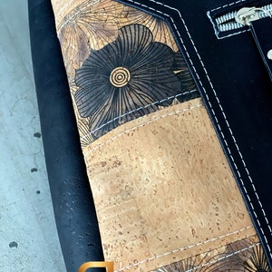 May include: Black leather bag with a cork and floral design. The bag has a zipper closure and white stitching.