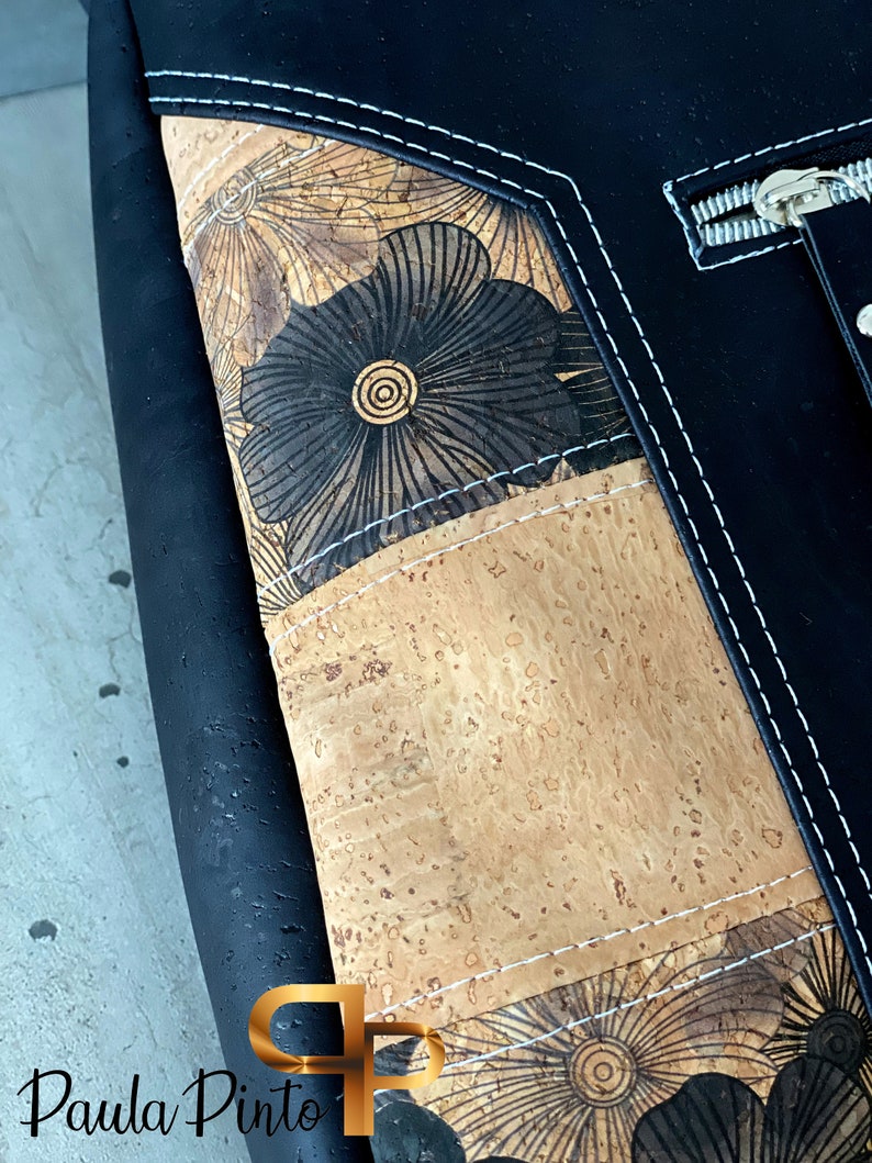 May include: Black leather bag with a cork and floral design. The bag has a zipper closure and white stitching.