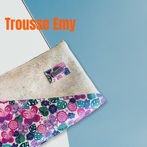 May include: A beige cork fabric makeup bag with a floral print on the bottom half. The bag has a silver zipper pull and a small silver tag with the word "Emy" on it. The text "Trousse Emy" is written in orange at the top of the image.