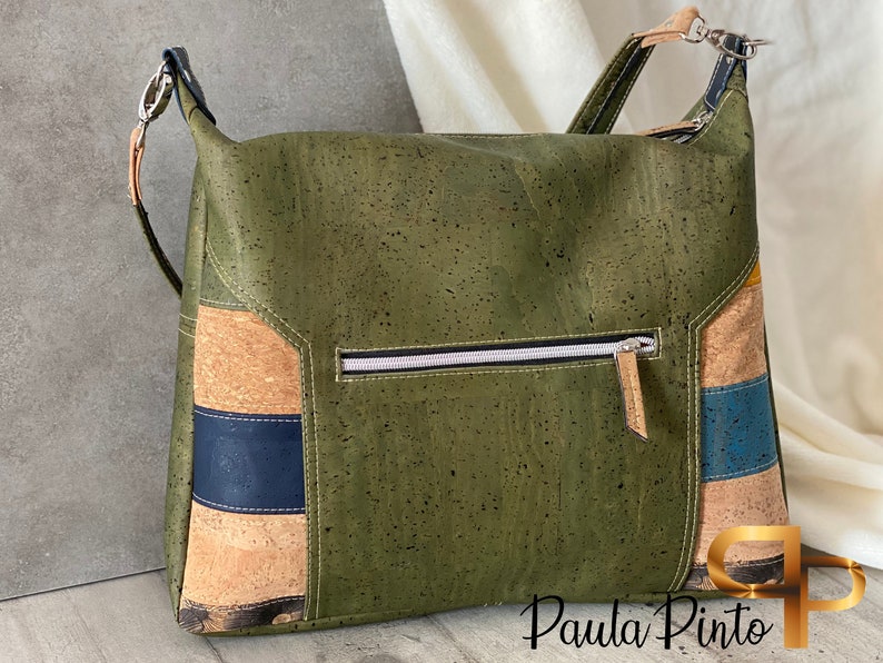 May include: A green cork handbag with a zipped pocket and a brown leather strap. The bag has a patchwork design with brown, blue and yellow accents. The text "Paula Pinto" is visible on the bottom right corner of the bag.